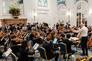 Junge Musiker spielen im Orchester unter der Leitung eines Dirigenten in elegantem Saal.